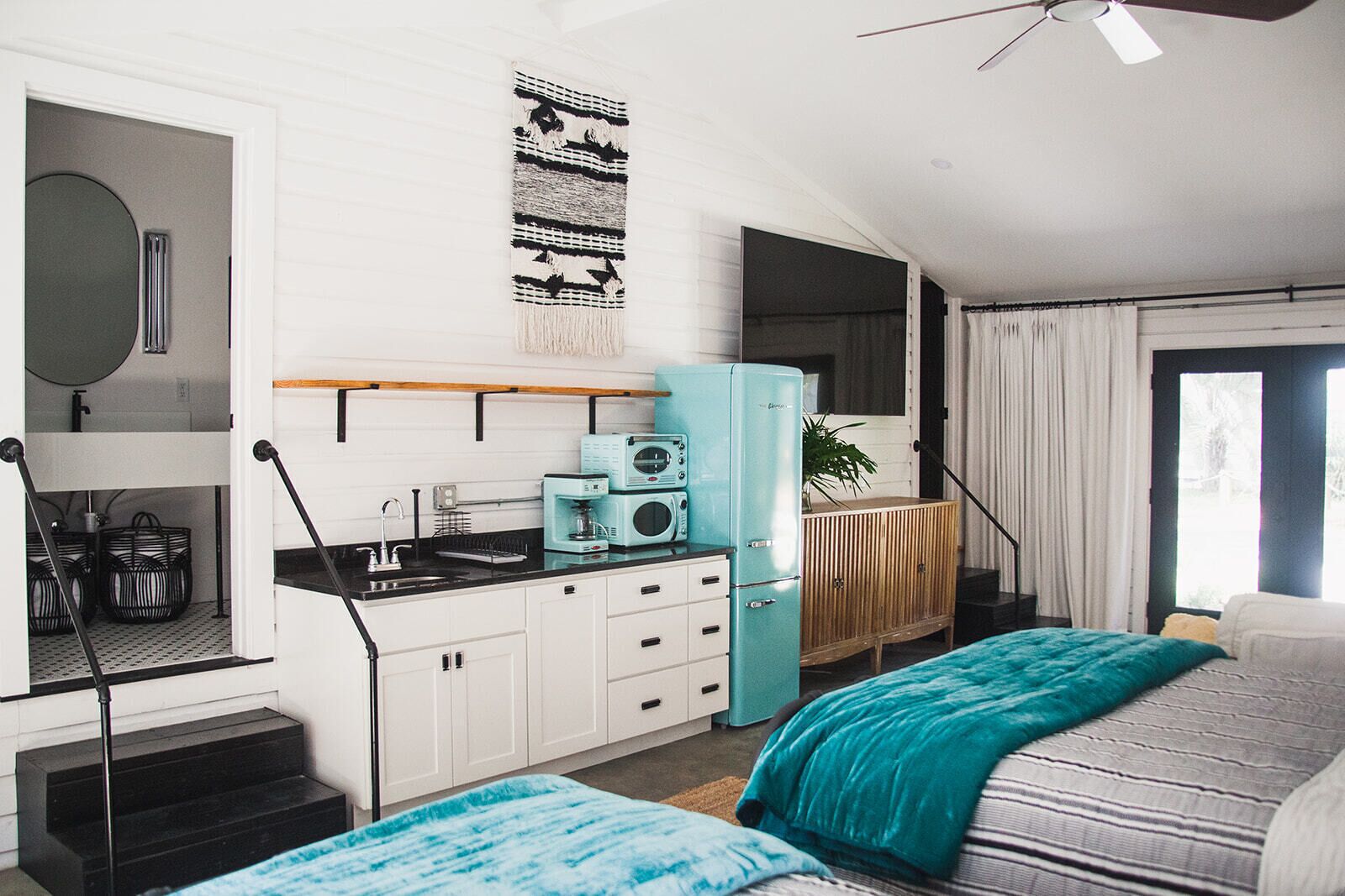 A compact, modern room with a kitchenette, a bed with a teal blanket, and a large window with curtains.