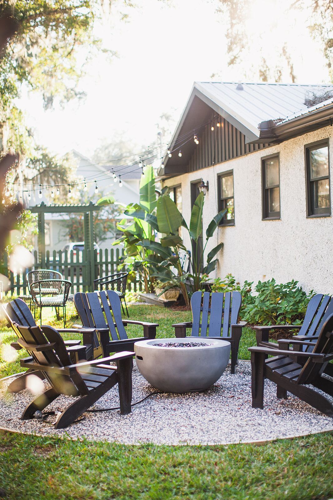 A patio with four Adirondack chairs, a fire pit, and a house in the background.