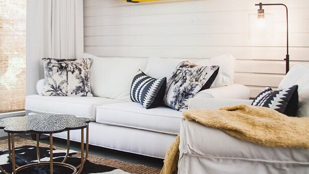 Ein modernes Wohnzimmer mit einem weißen Sofa, gemusterten Kissen, einem runden Couchtisch und einem an der Wand befestigten Surfbrett.