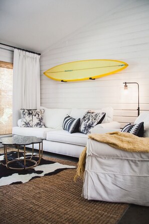 Un salon moderne avec un canapé blanc, des coussins à motifs, une table basse ronde et une planche de surf accrochée au mur.