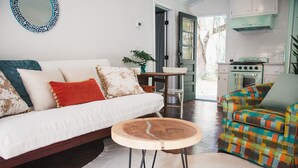 A cozy living room with a sofa, a colorful armchair, a round wooden coffee table, and a kitchen area with a stove.