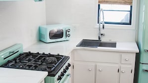 A compact kitchen with white cabinets, a teal stove, a small window with blinds, and a hanging lantern.