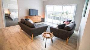 Ein modernes Wohnzimmer mit zwei Sofas, einem Couchtisch und einem Fernseher auf einer Holzkommode. Durch das große Fenster ist ein Balkon mit Geländer zu sehen.
