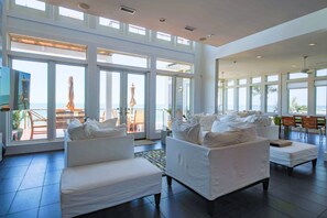 Een ruime woonkamer met grote ramen, uitzicht op de oceaan en een zithoek met witte banken en stoelen.