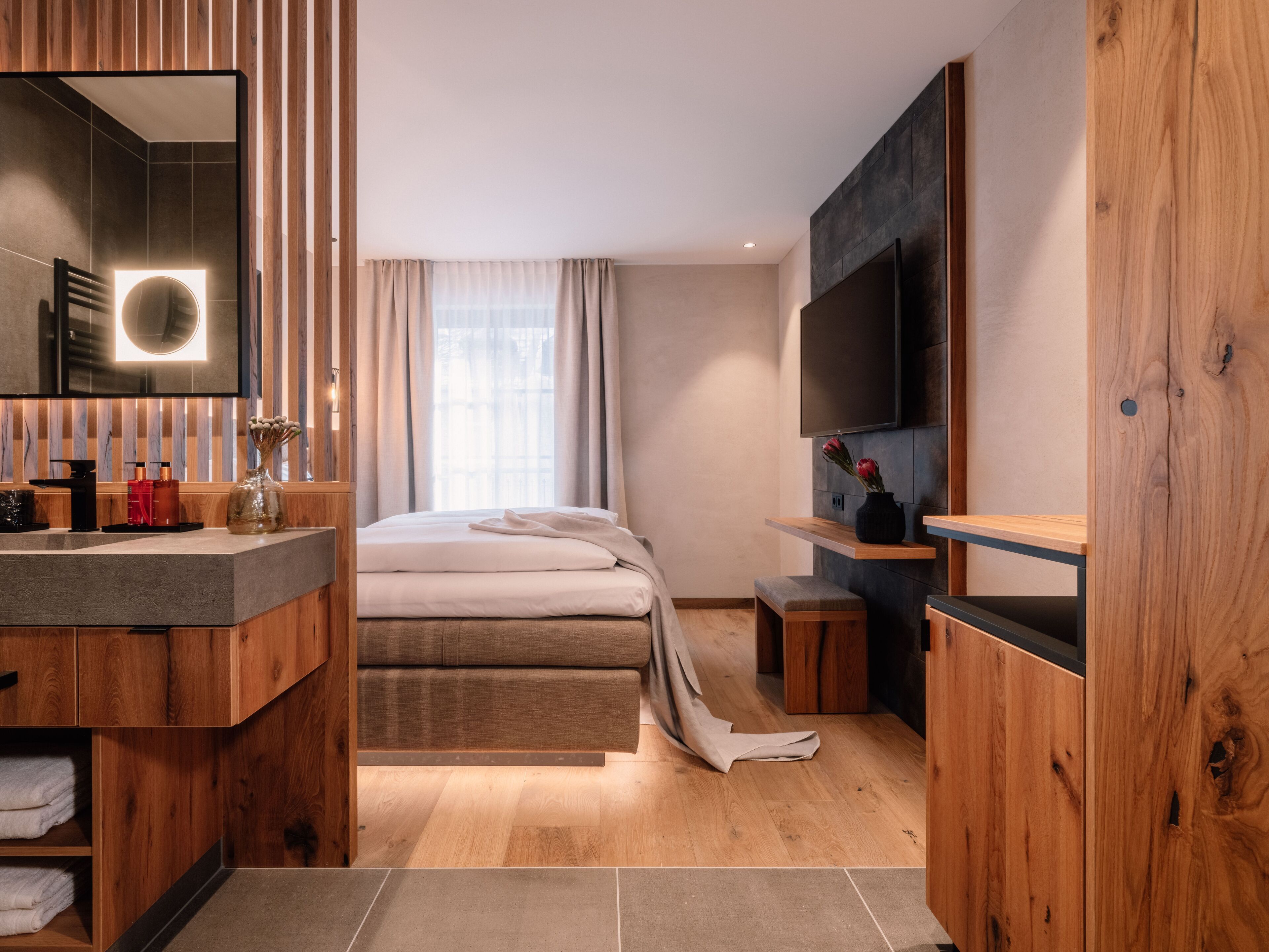 A modern hotel room with a wooden vanity, a large bed, a flat-screen TV, and a large window with curtains.