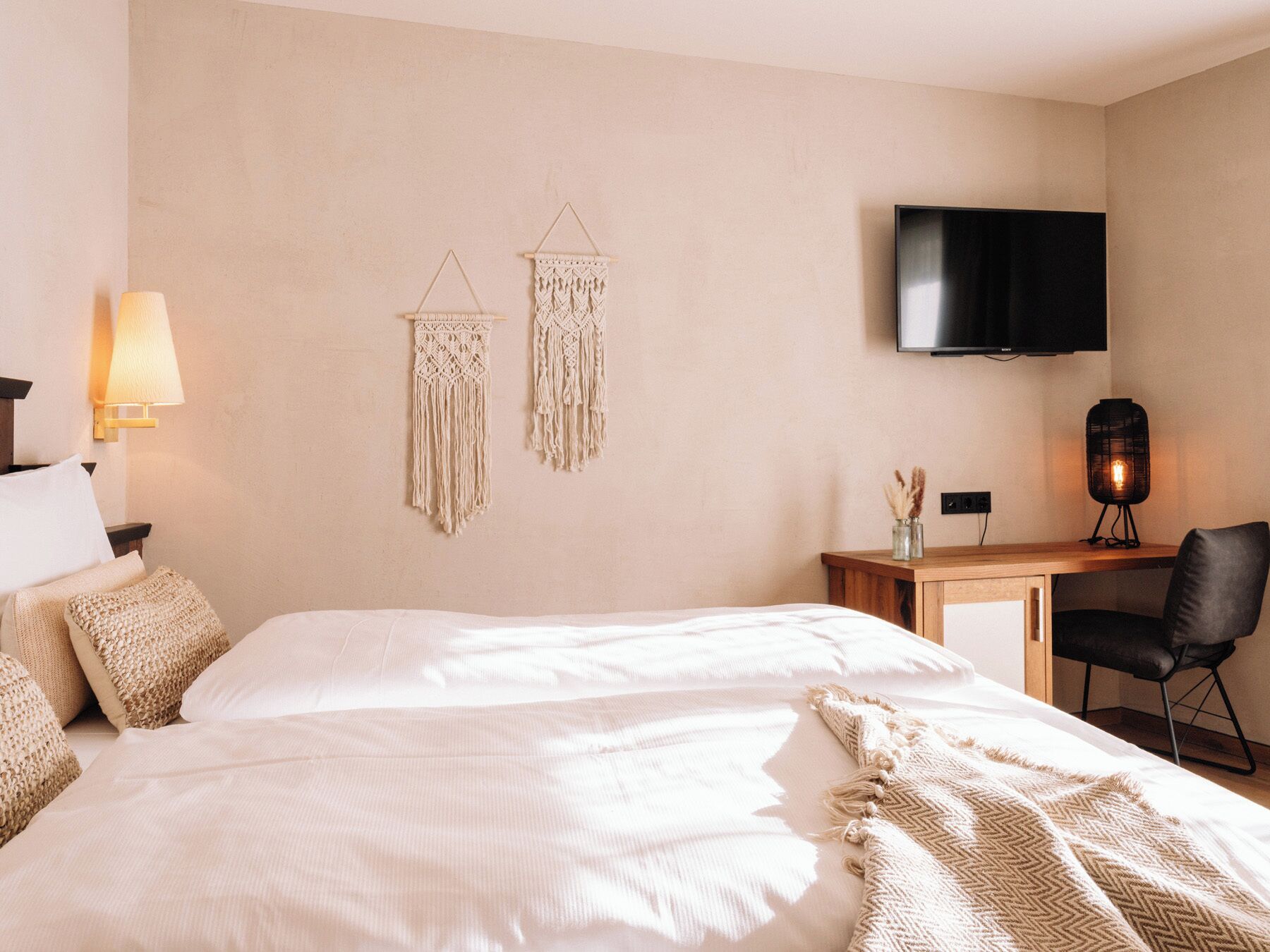 A hotel room with a large bed, a desk with a chair, a TV mounted on the wall, and two wall-mounted macramé decorations.