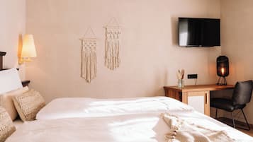A hotel room with a large bed, a desk with a chair, a TV mounted on the wall, and two wall-mounted macramé decorations.