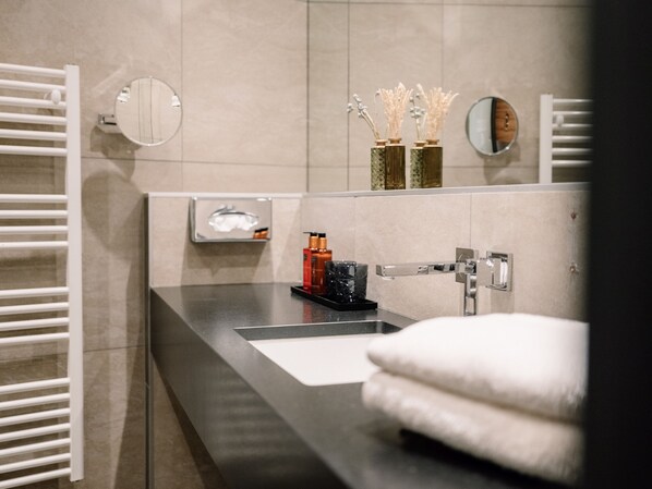 Un baño moderno con encimera negra, lavabo blanco y espejo. Hay dos jarrones con flores blancas sobre la encimera y un perchero con una toalla blanca.