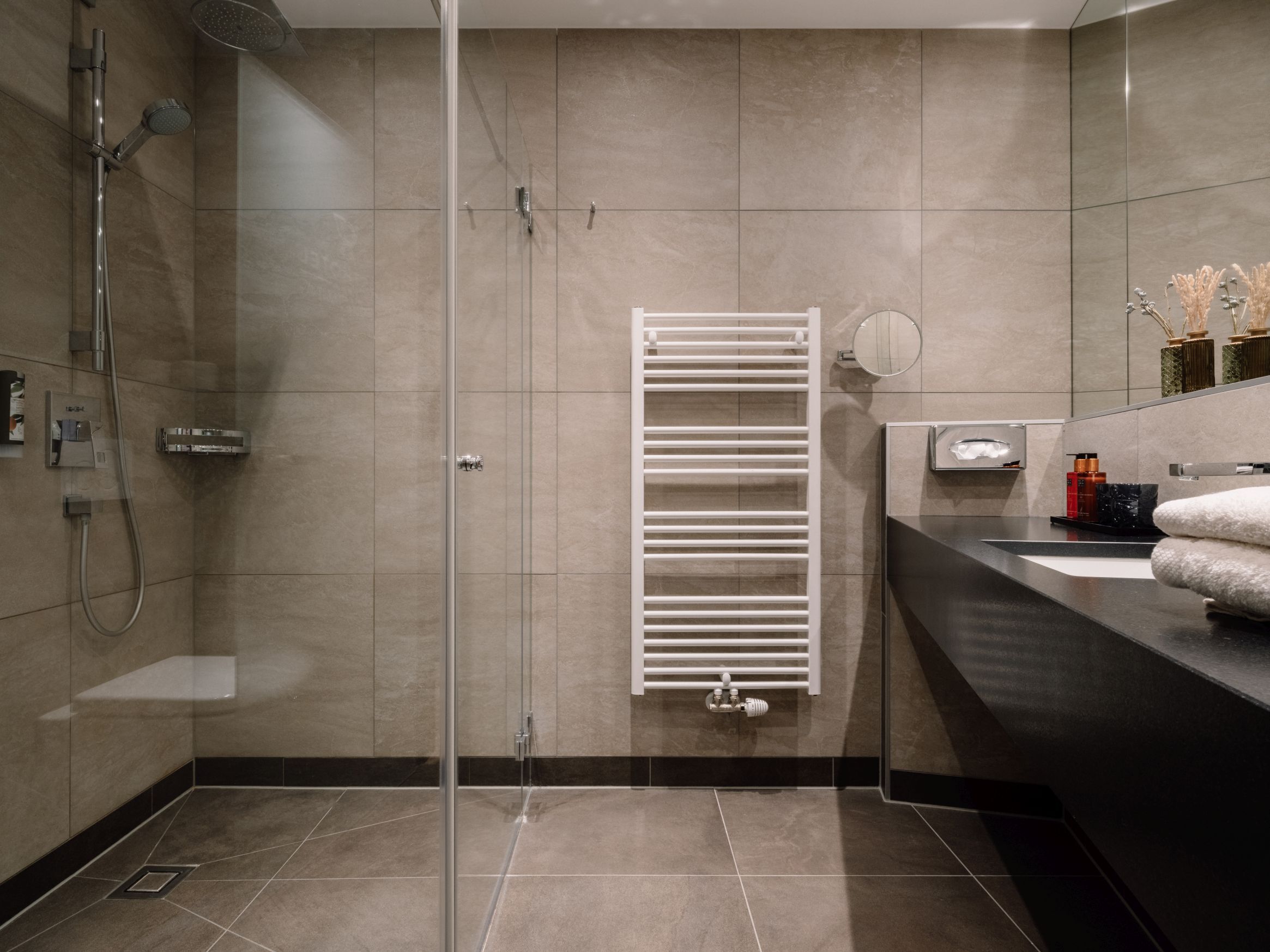 Casa de banho moderna com cabine de duche em vidro, toalheiro branco e bancada preta com lavatório e produtos de higiene pessoal.