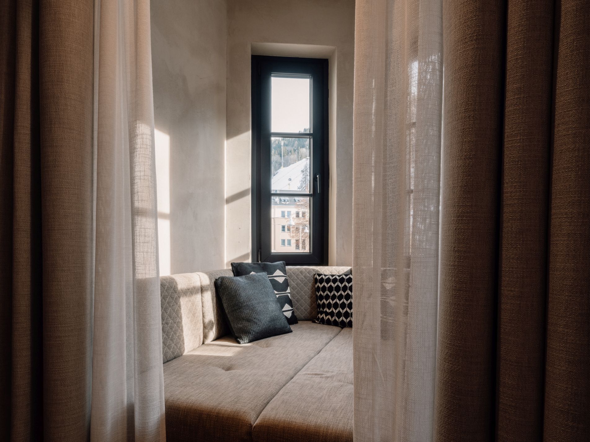 Une pièce chaleureuse avec une fenêtre donnant sur un paysage enneigé, un banc rembourré agrémenté de coussins à motifs et de rideaux légers.