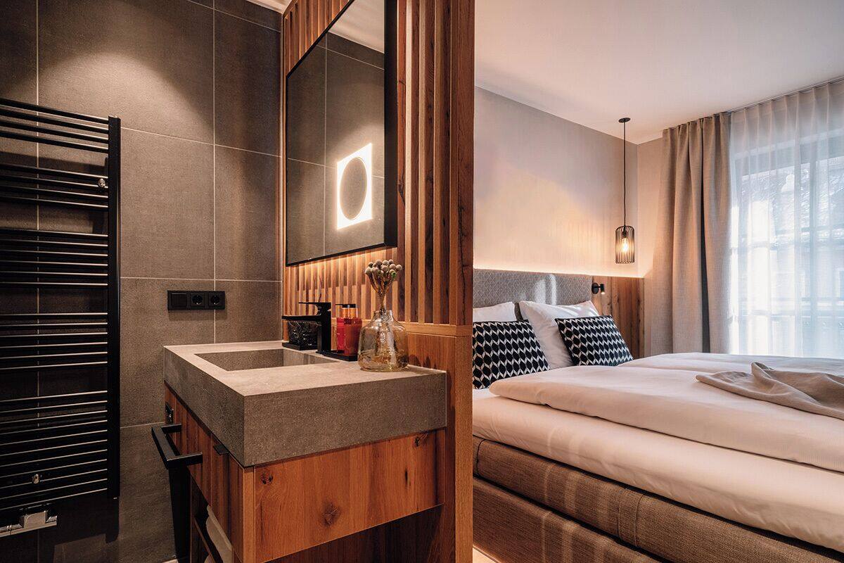 Une salle de bain moderne avec un meuble-lavabo en bois, un grand miroir et un radiateur encastré. La pièce comprend un grand lit avec une tête de lit, une table de chevet et un luminaire suspendu.