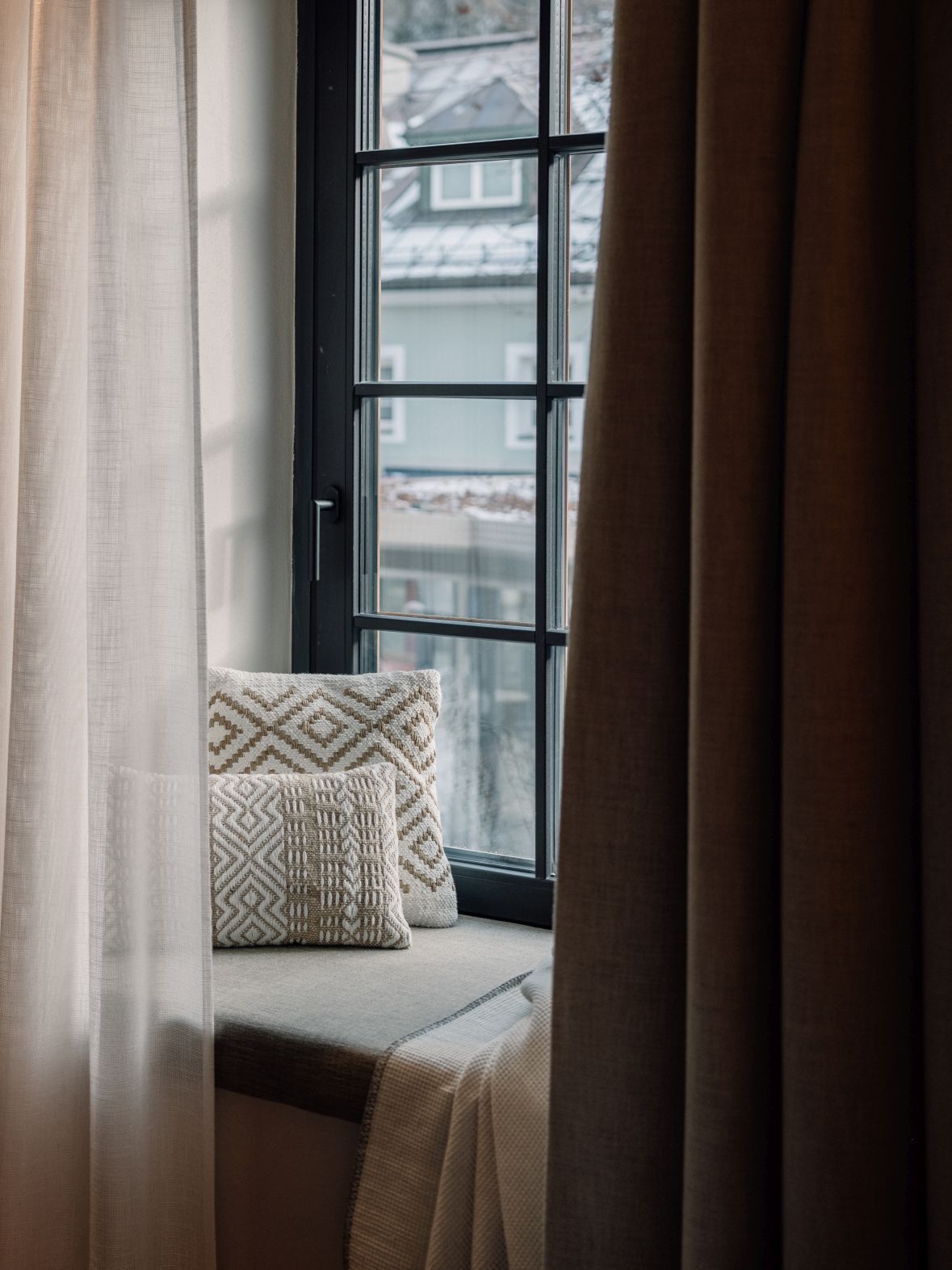 Ein Fensterplatz mit gemustertem Kissen und Decke, teilweise gezogene Vorhänge und Blick auf Gebäude.