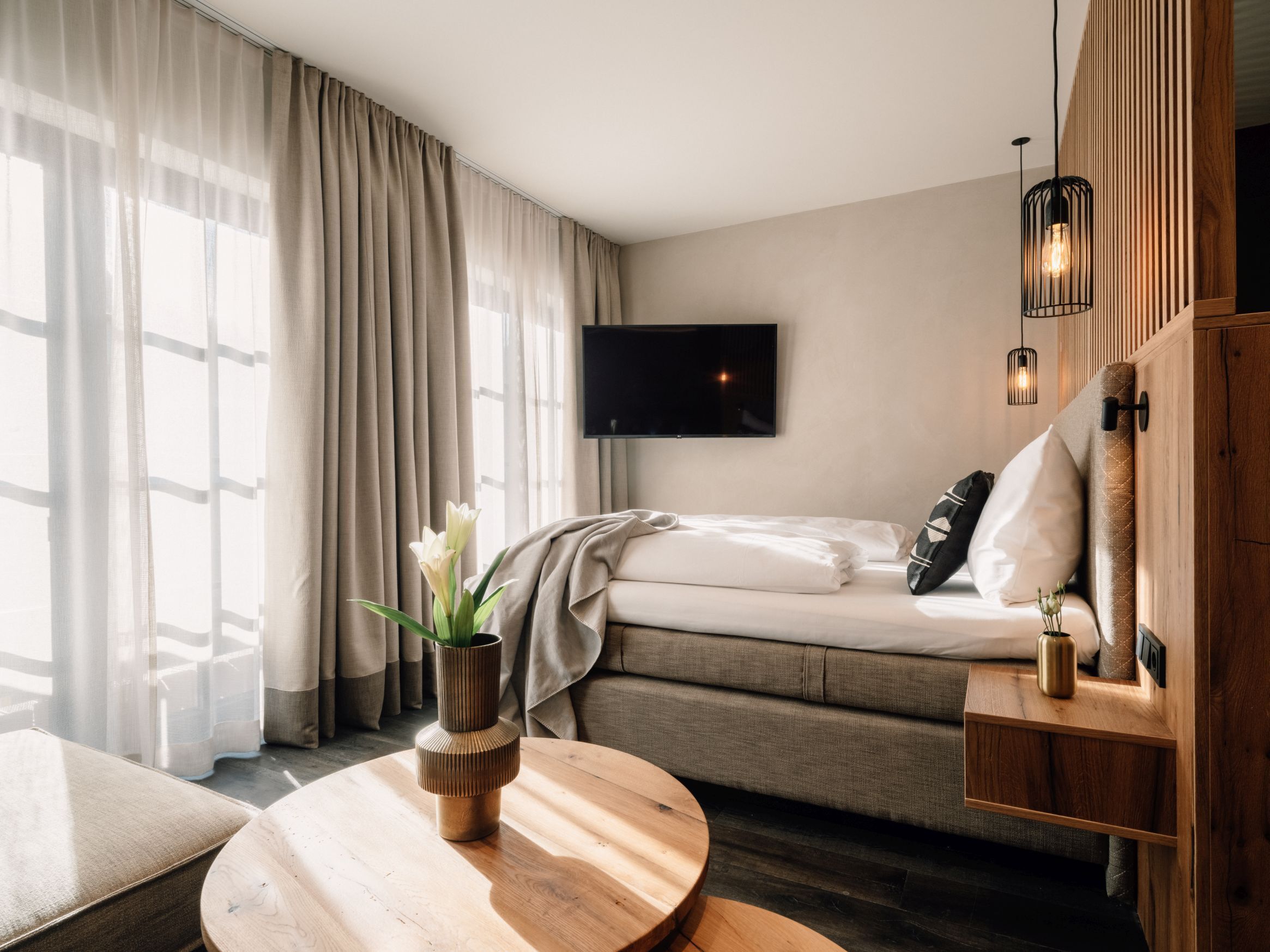 A modern hotel room with a sofa bed, a flat-screen TV, a wooden side table, and a round wooden table.