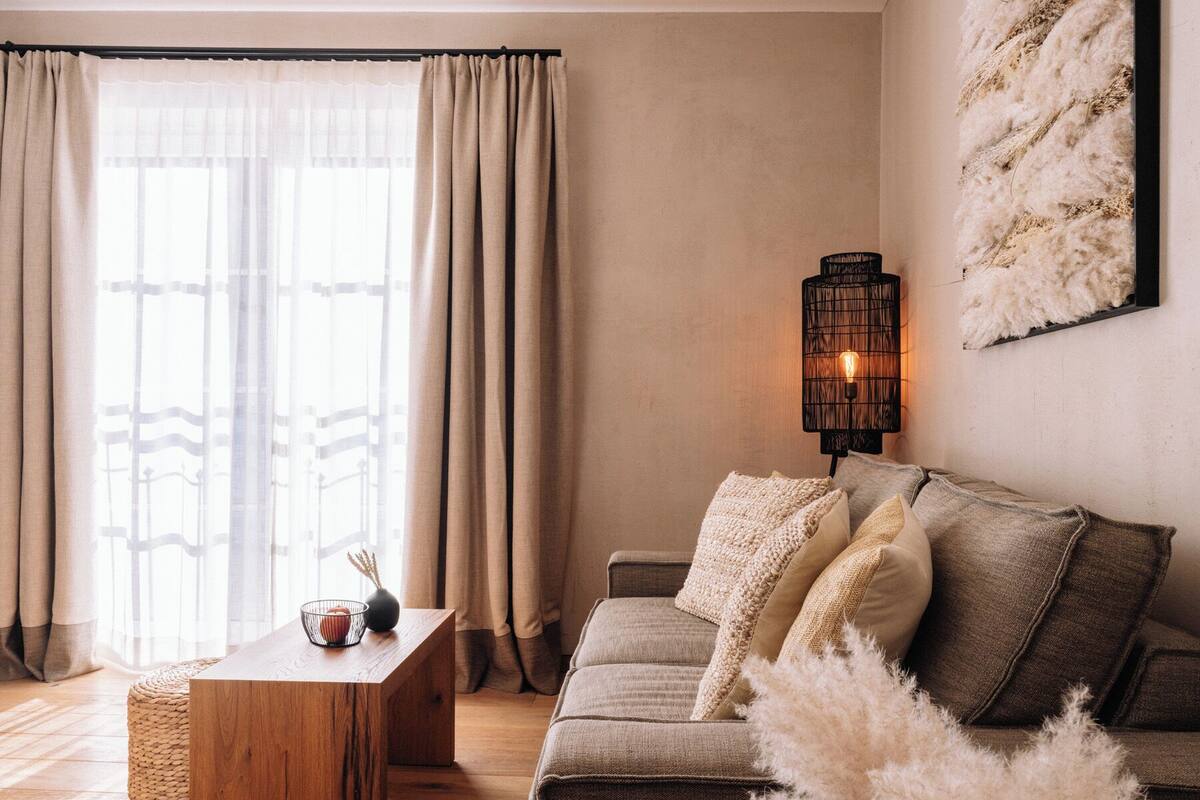 Un salon chaleureux avec une table basse en bois, un canapé agrémenté de coussins, une lampe fixée au mur et une grande fenêtre avec des rideaux.