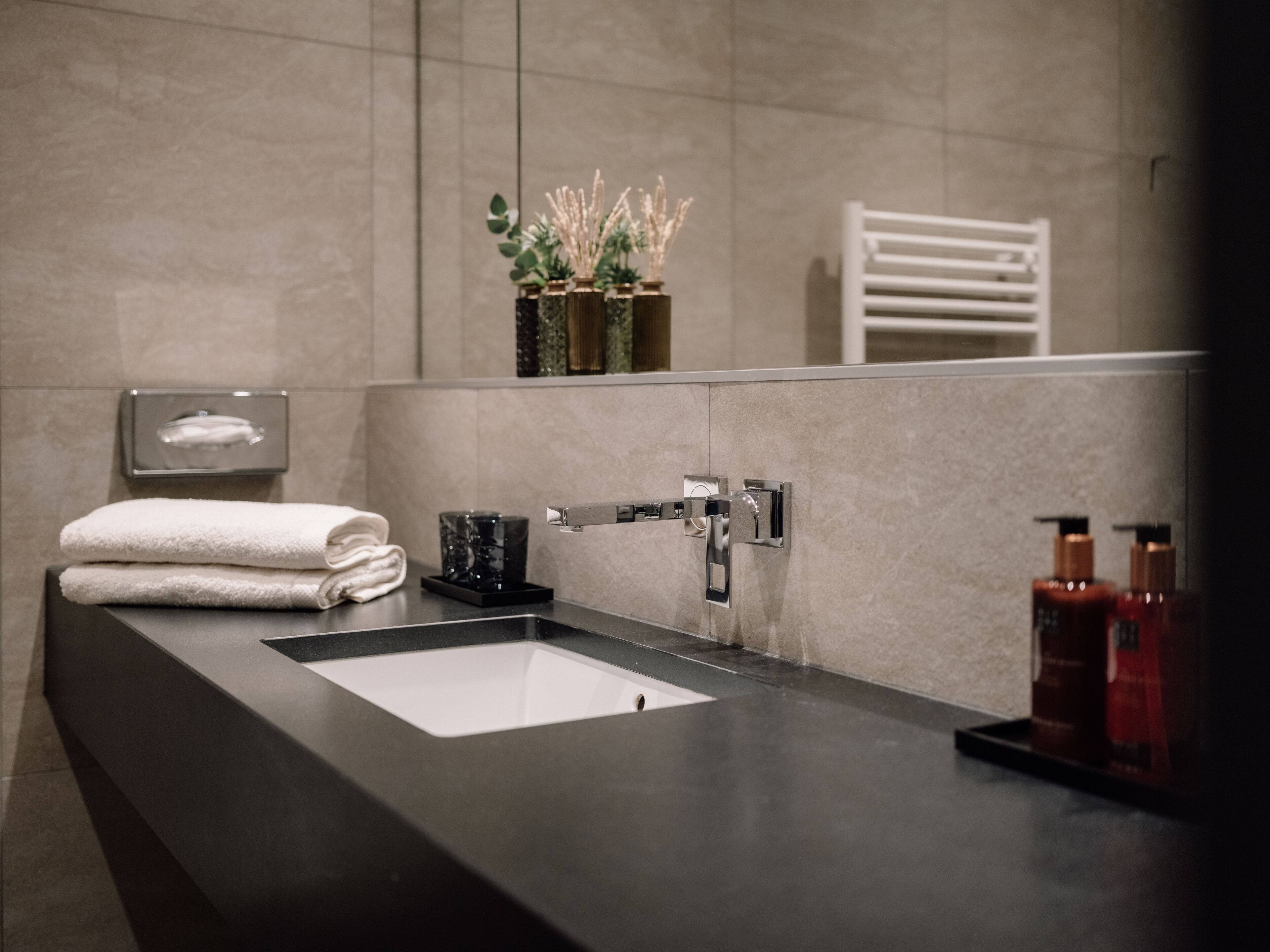 A modern bathroom with a large mirror, a black countertop, a white sink, and a towel rack.
