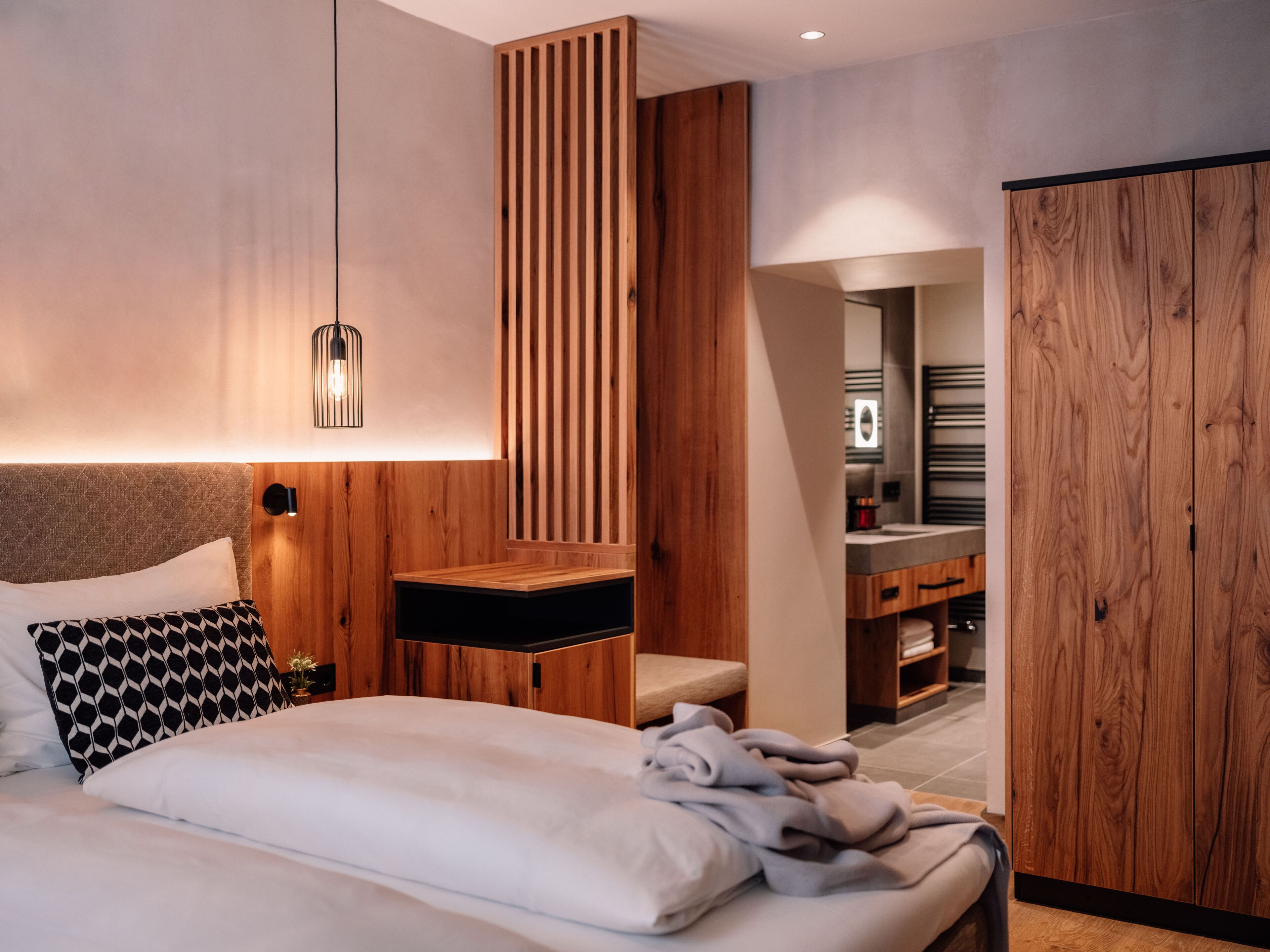 Un dormitorio moderno con cabecera de madera, una mesita de noche, una lámpara colgante y vistas al baño.