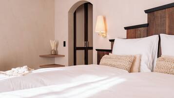 Una cama bien hecha con sábanas blancas y dos almohadas con textura, una lámpara de noche y una mesita de madera con un jarrón de flores secas.