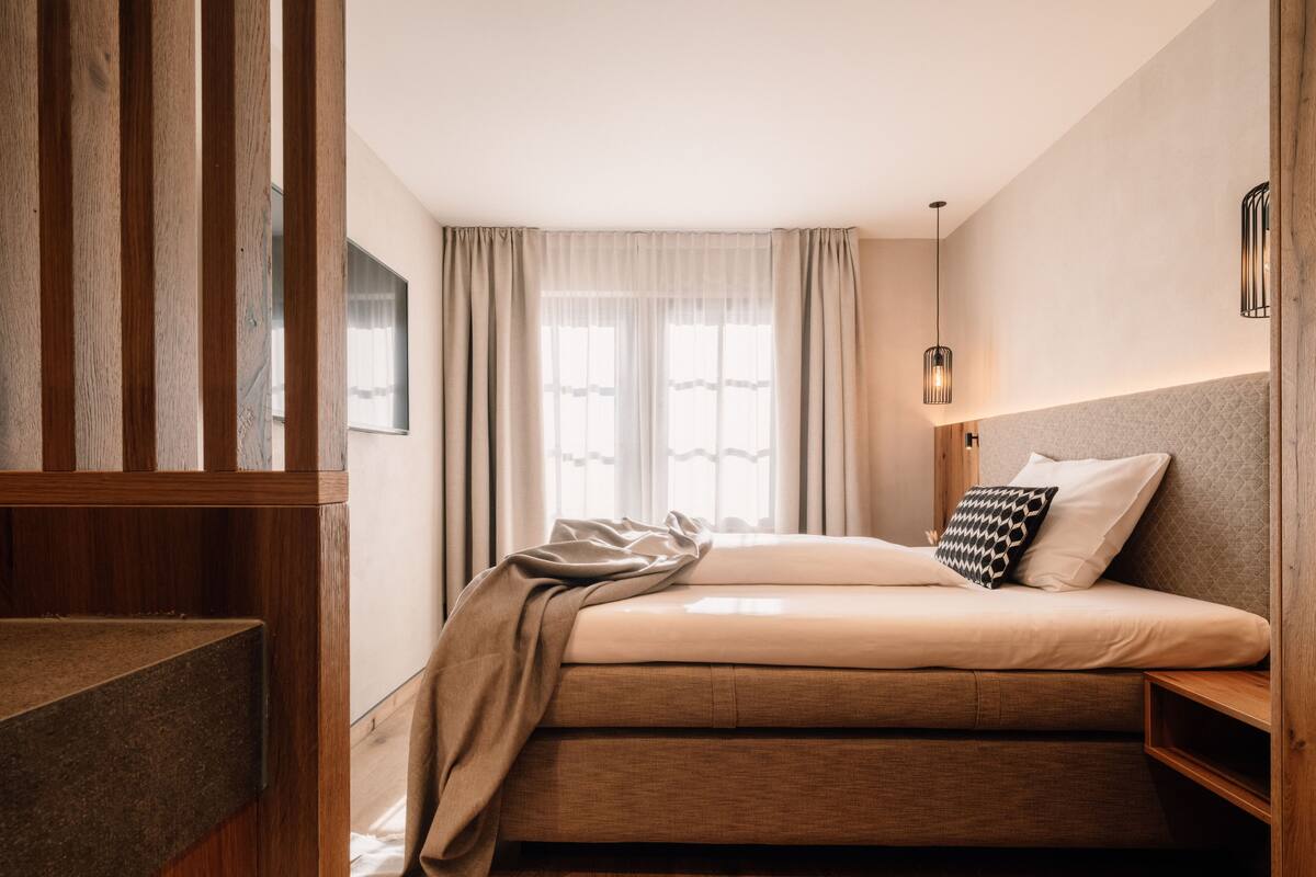 Une chambre à coucher avec un lit, une tête de lit en bois, une table de chevet, un luminaire suspendu et une fenêtre avec des rideaux.