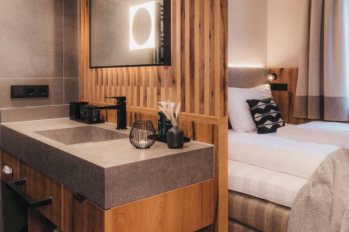 Une chambre d’hôtel moderne avec une coiffeuse en bois, un grand miroir, un lit avec du linge de maison blanc et une fenêtre avec des rideaux.