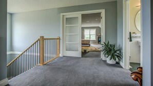 Un pasillo con suelo de alfombra, una escalera de madera y una puerta que da a una habitación con cama y ventana.