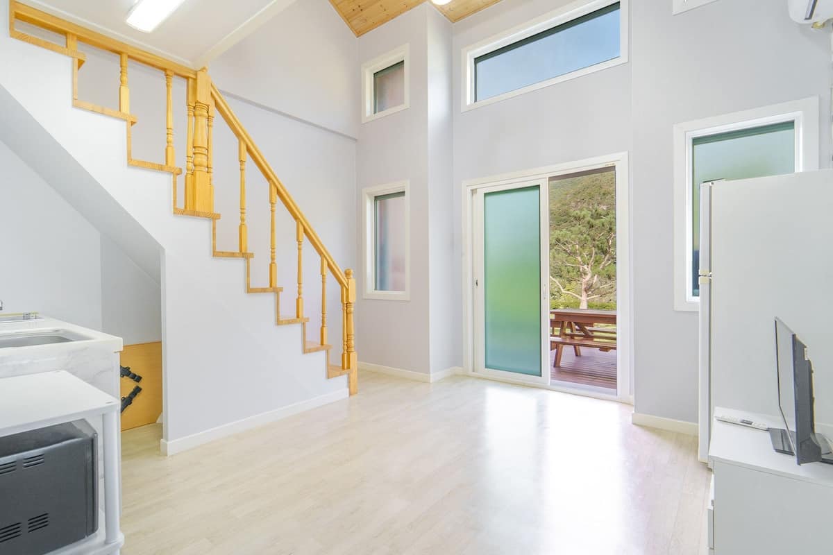 A spacious room with a wooden staircase, a kitchen area, and a sliding glass door leading to an outdoor deck.