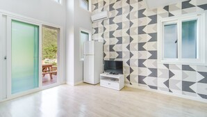 Un appartamento moderno con una parete decorata da un motivo geometrico, una televisione a schermo piatto su un supporto bianco, un frigorifero e una porta scorrevole in vetro che dà su una zona esterna con un tavolo di legno.