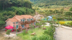 Una casa a due piani con un balcone, circondata da un giardino curato e un sentiero sterrato che conduce a una serra blu.