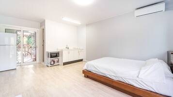 Ein modernes Schlafzimmer mit einem hölzernen Bett, einem weißen Kleiderschrank, einer kleinen Küchenzeile und einer Glastür, die auf einen Balkon führt.