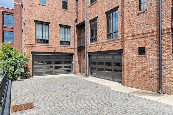 A brick building with two garage doors, a paved courtyard, and a metal fence.