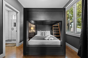 A modern bedroom with a bunk bed, a bedside table with a lamp, a wooden ladder, and a view of the outdoors through a window.