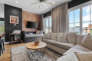 A modern living room with a sectional sofa, a coffee table, a TV mounted on a brick wall, and large windows with curtains.