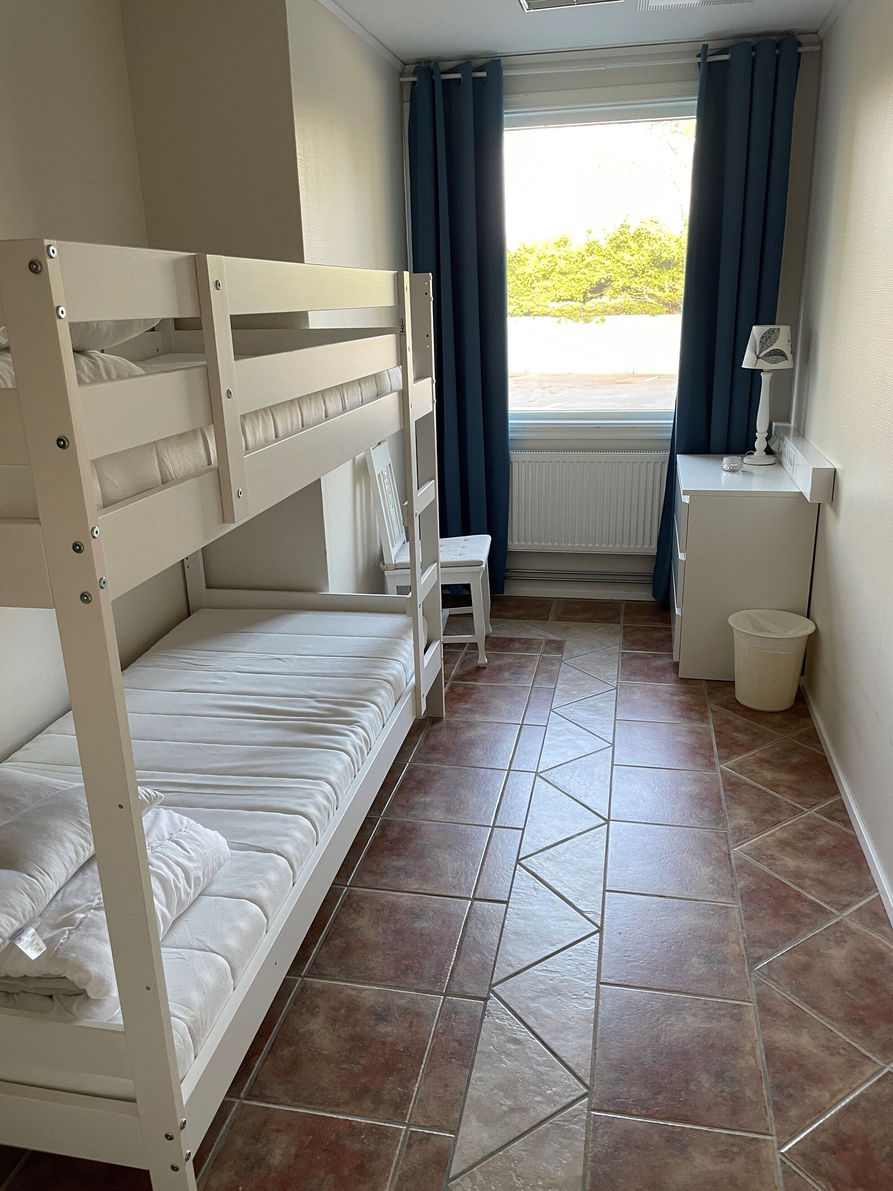 A small, clean room with bunk beds, a desk, a chair, and a window with curtains.