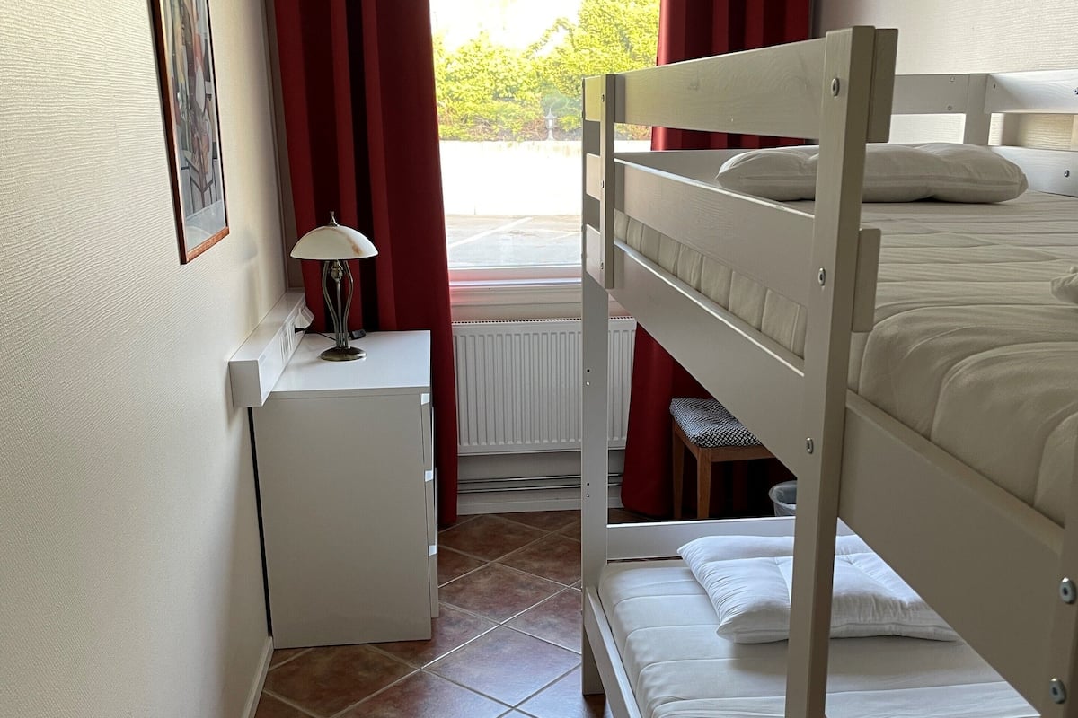 Una habitación pequeña y bien iluminada con una cama litera, una mesita de noche con lámpara, un radiador y una ventana con cortinas rojas.