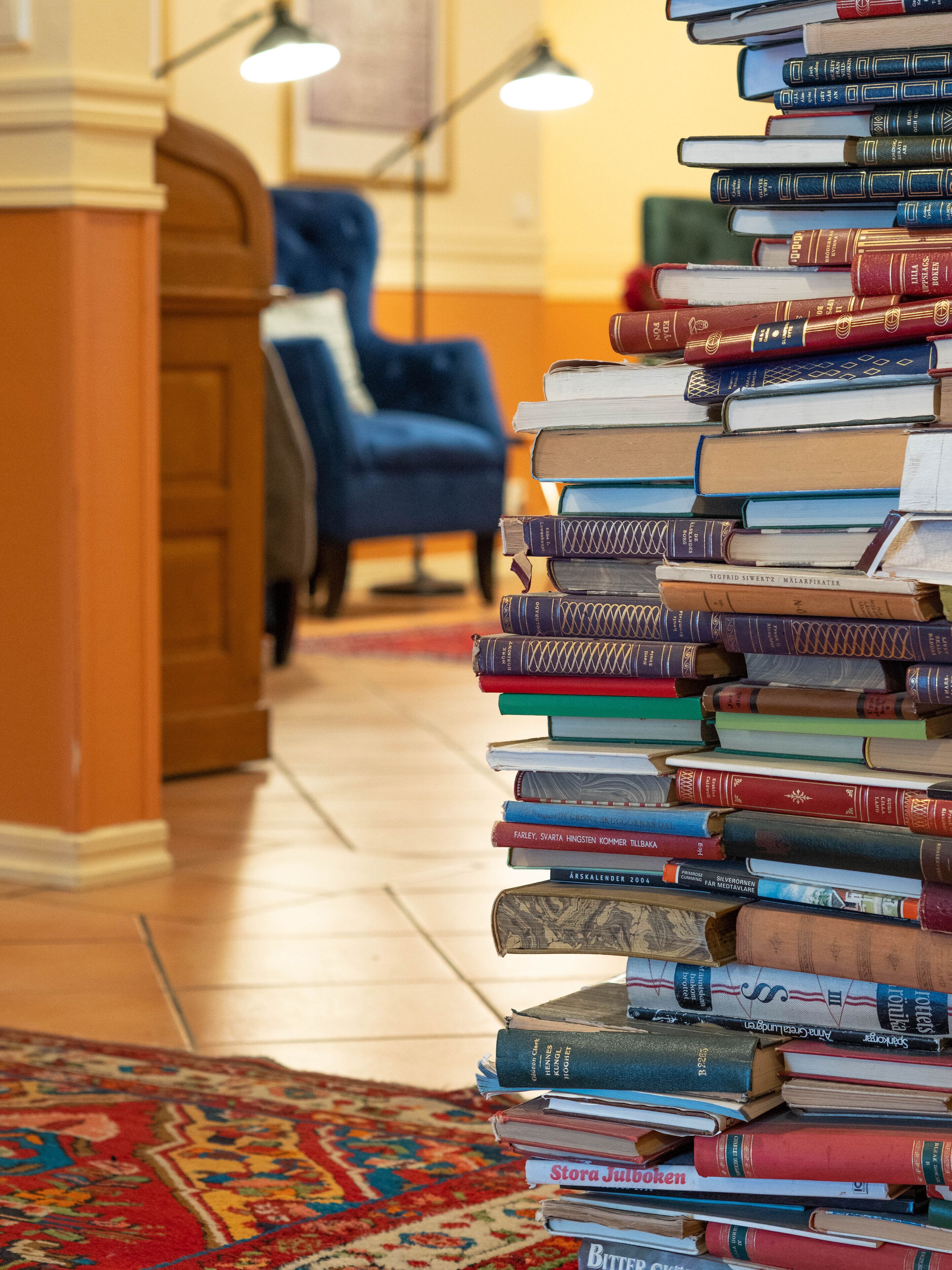Ein Bücherstapel in einem Raum mit blauem Sessel und einem gemusterten Teppich.