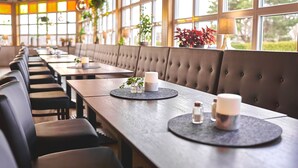 A restaurant with long wooden tables, cushioned booths, and large windows with plants. - Björkbackens Vandrarhem i Vimmerby - Hostel (Vimmerby)