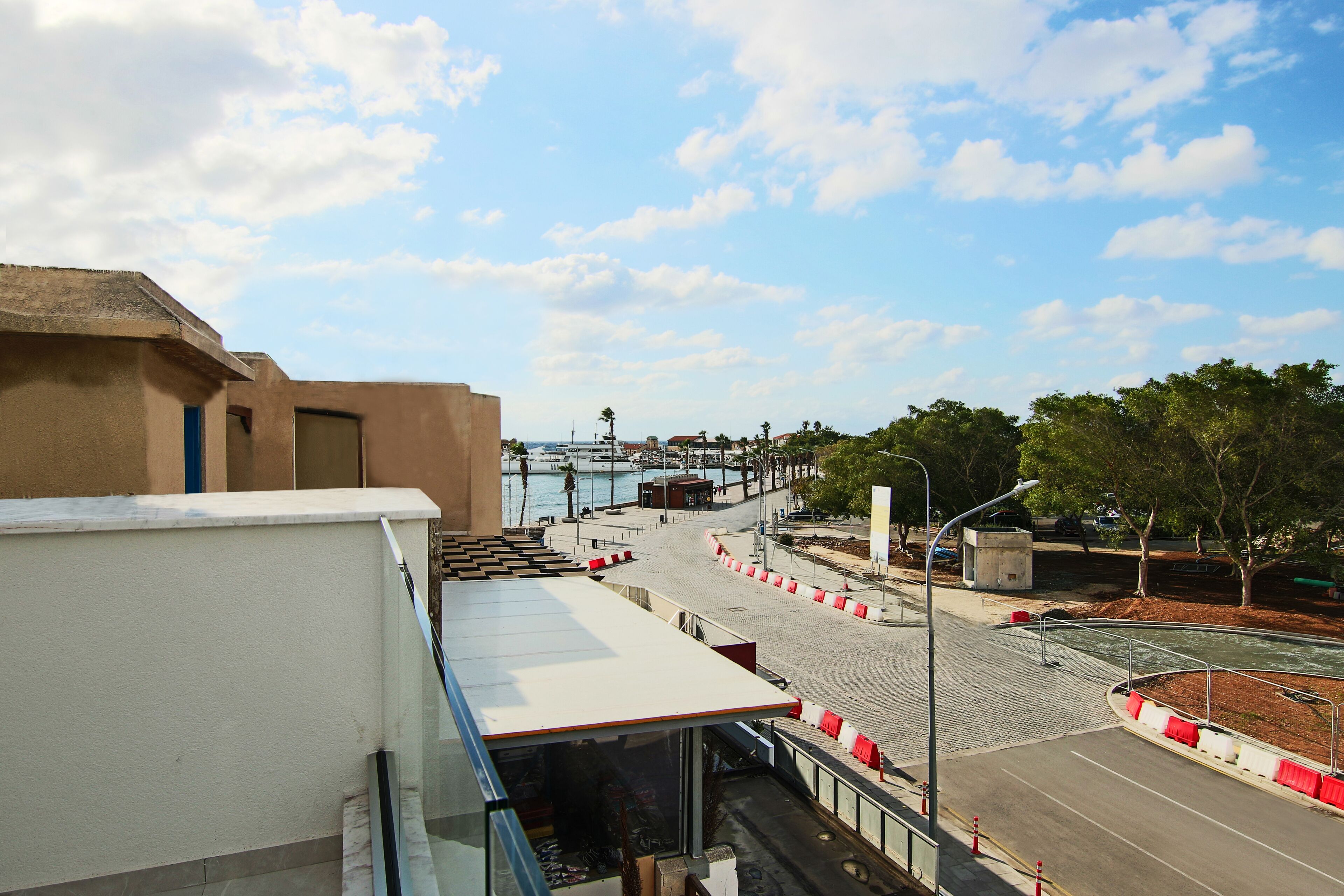 Vista costeira com uma estrada que leva a um porto de iates, um calçadão e um edifício com telhado plano.