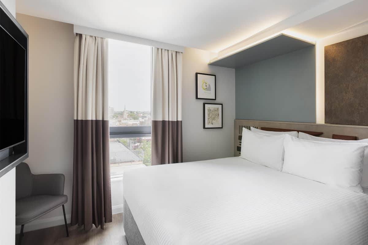 A hotel room with a large bed, a TV, a chair, and a window with curtains.
