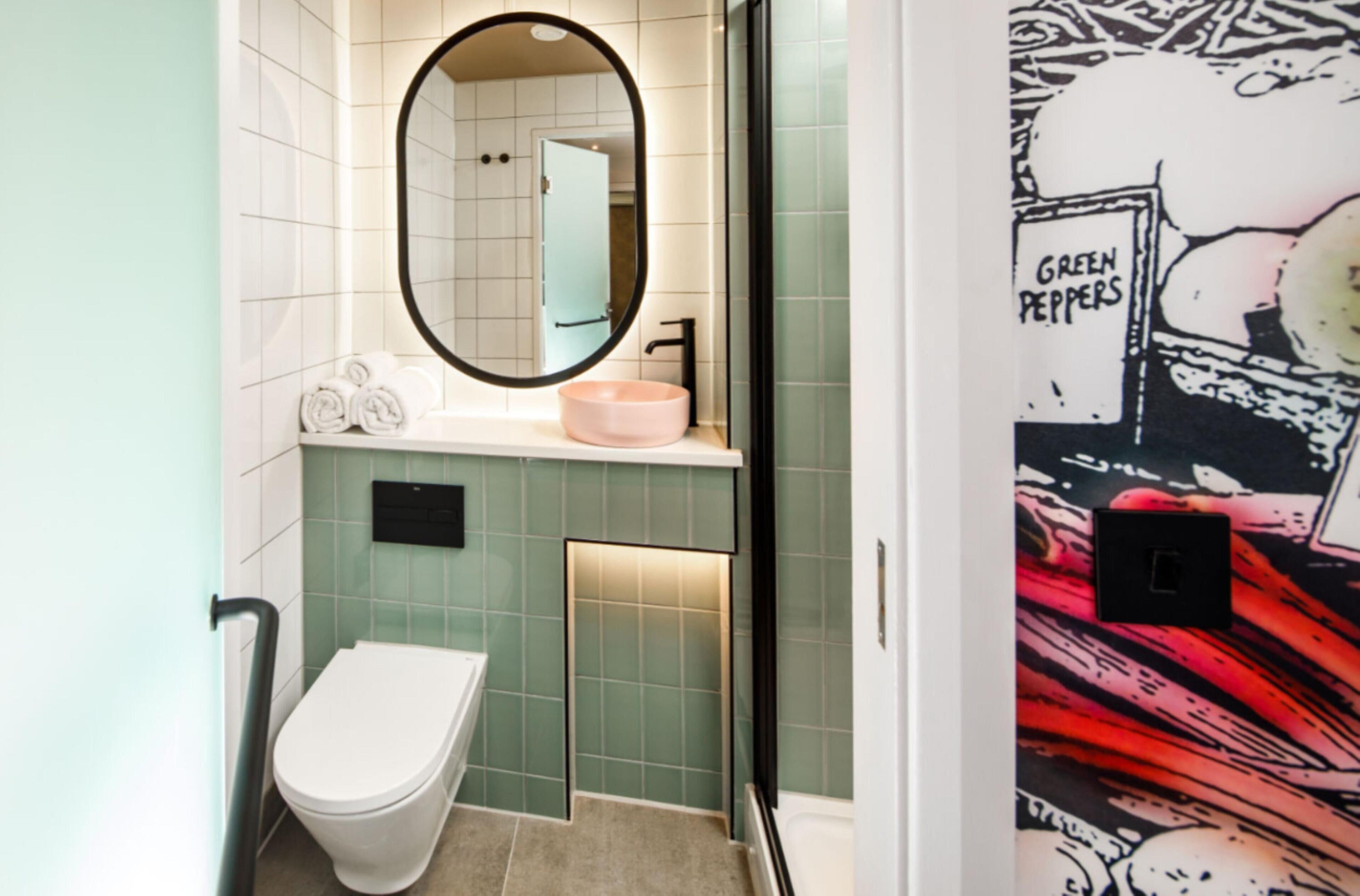 Un baño con inodoro blanco, espejo redondo, lavamanos rosa y paredes con azulejos verdes.