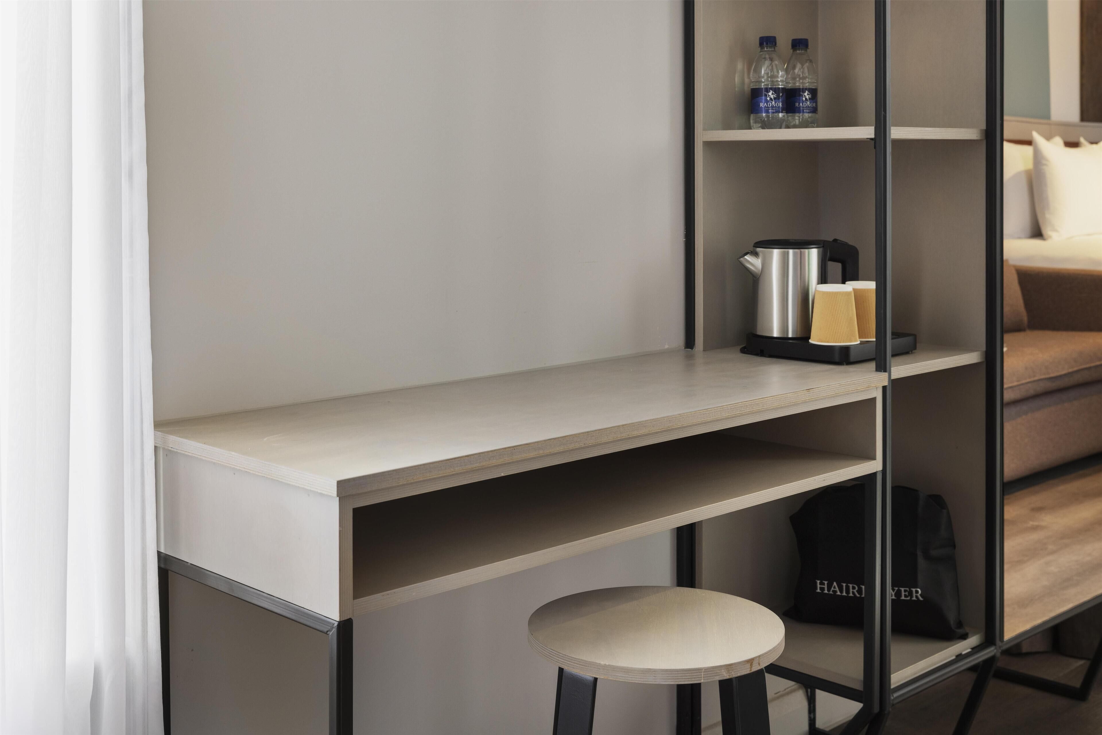 A modern hotel room with a minimalist design, featuring a wooden desk, a small stool, a shelf with toiletries, and a bed with a pillow.