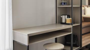 A modern hotel room with a minimalist design, featuring a wooden desk, a small stool, a shelf with toiletries, and a bed with a pillow.