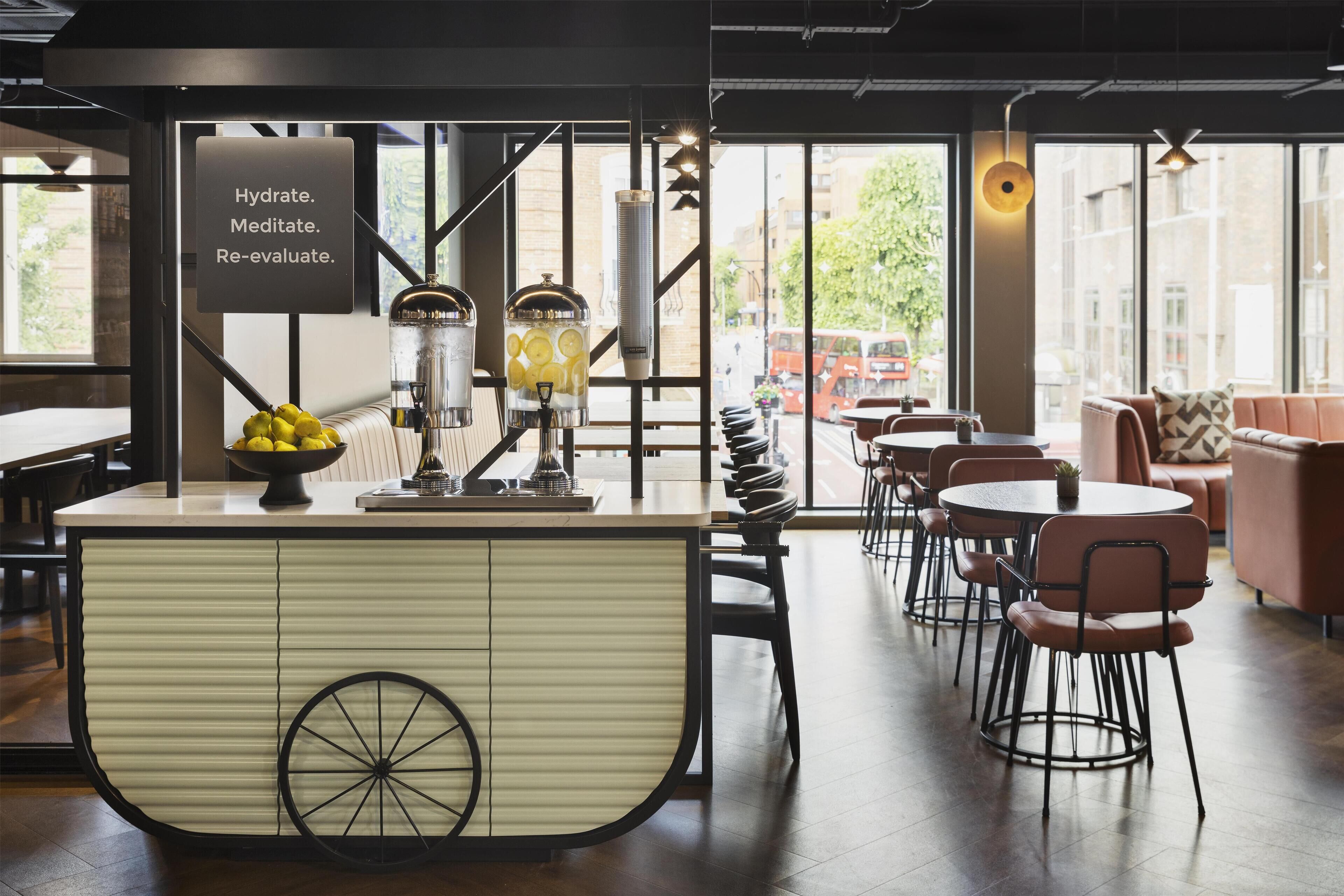 Un café moderne doté d’un bar central, comprenant un espace dédié à l’hydratation, des coins salon et de grandes fenêtres offrant une vue sur la ville.