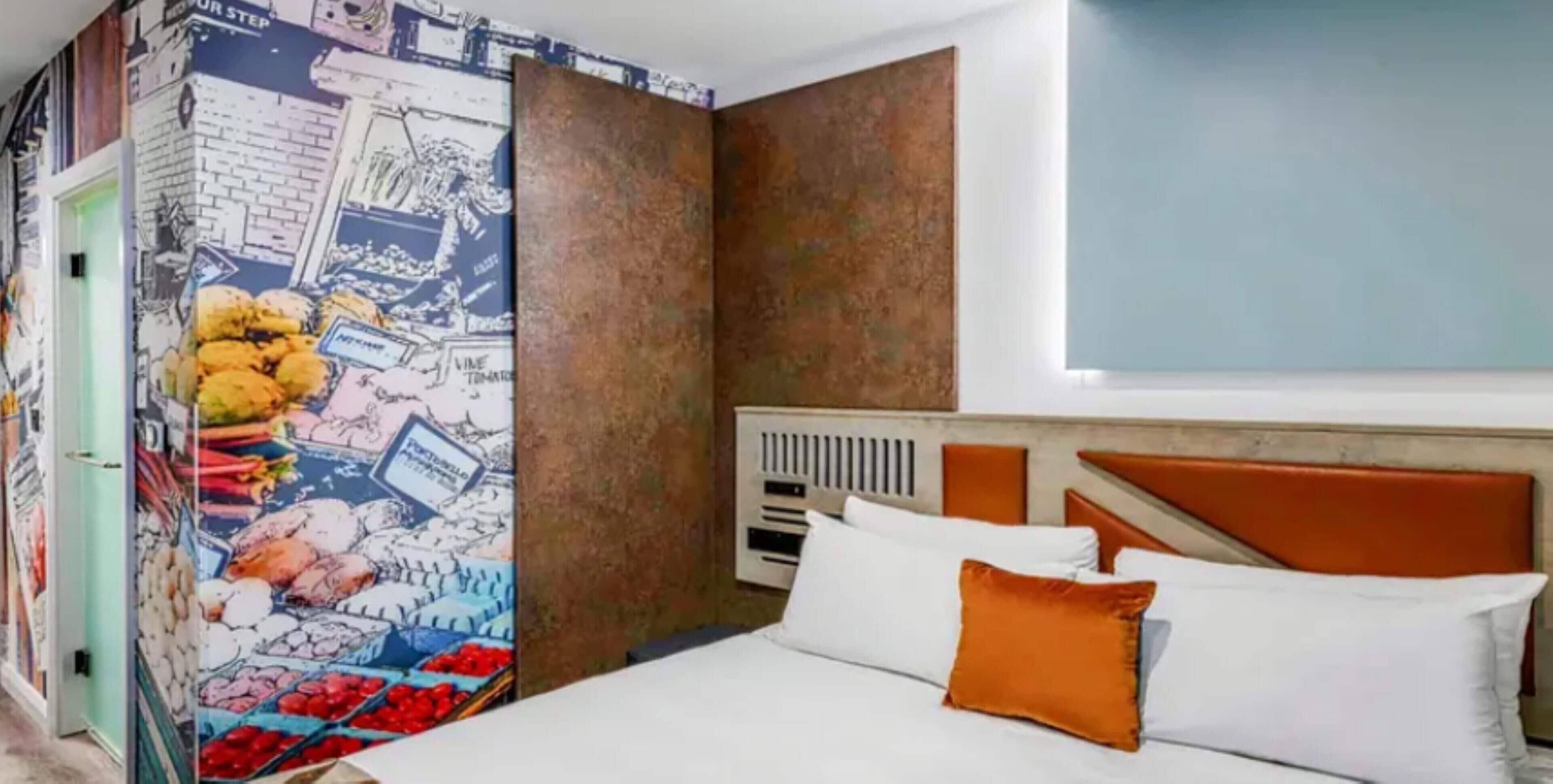 A hotel room with a bed, orange headboard, white bedding, and a mural of a market scene on the wall.