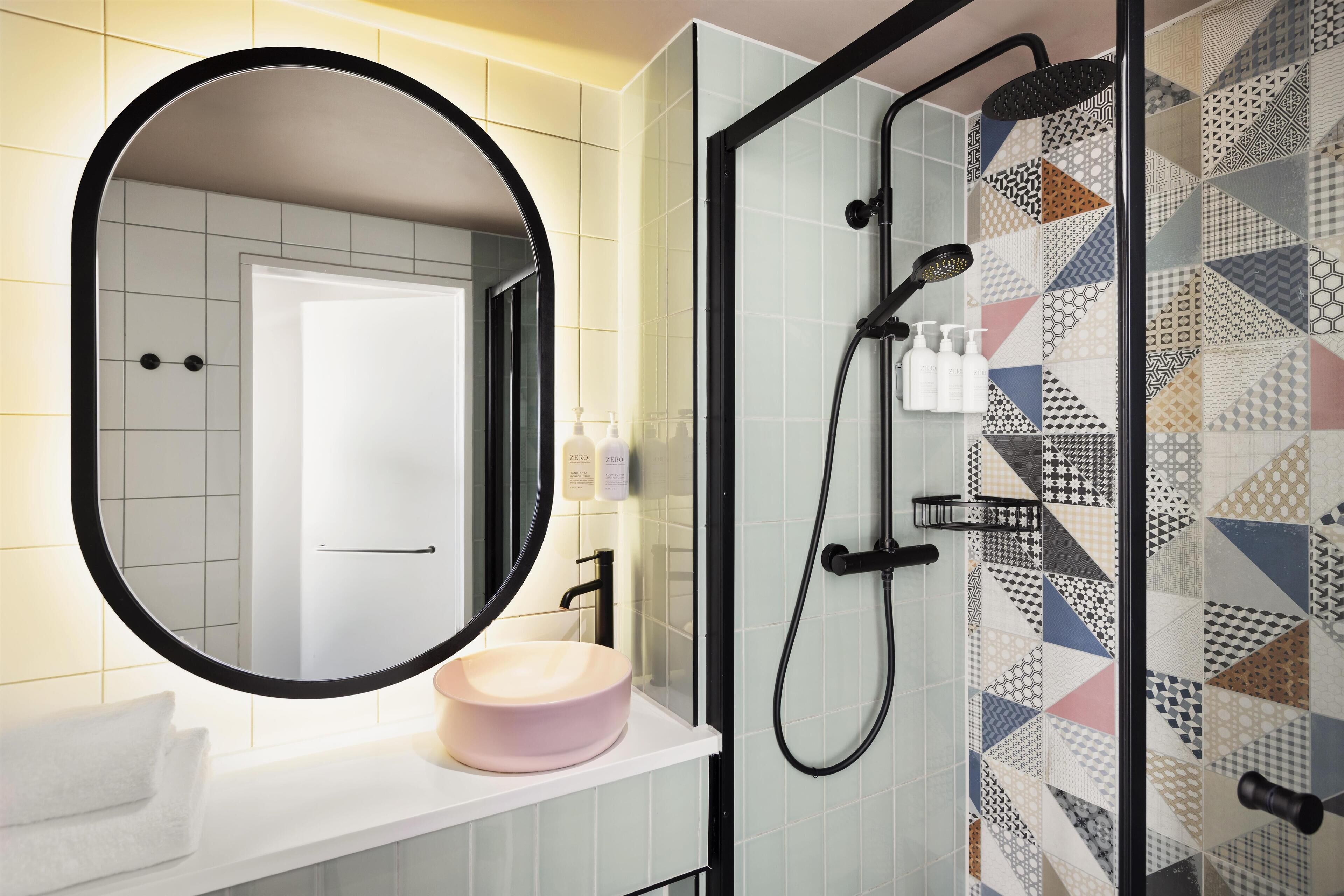 Un baño con un espejo redondo, un lavabo con una pileta color beige y una ducha con un diseño de azulejos geométricos.