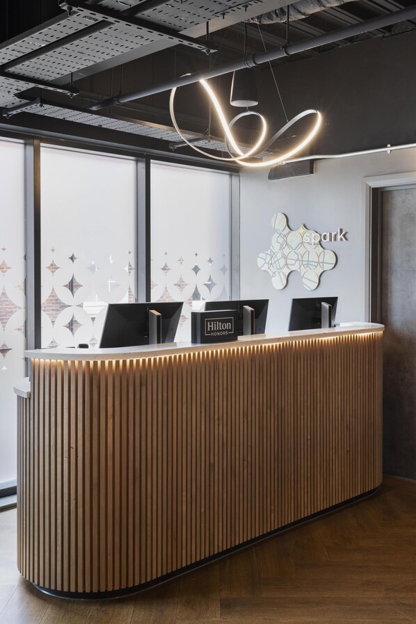 A modern hotel reception desk with a wooden finish, two black monitors, and a 'Hilton Honors' sign. - Spark by Hilton London Romford (Romford)