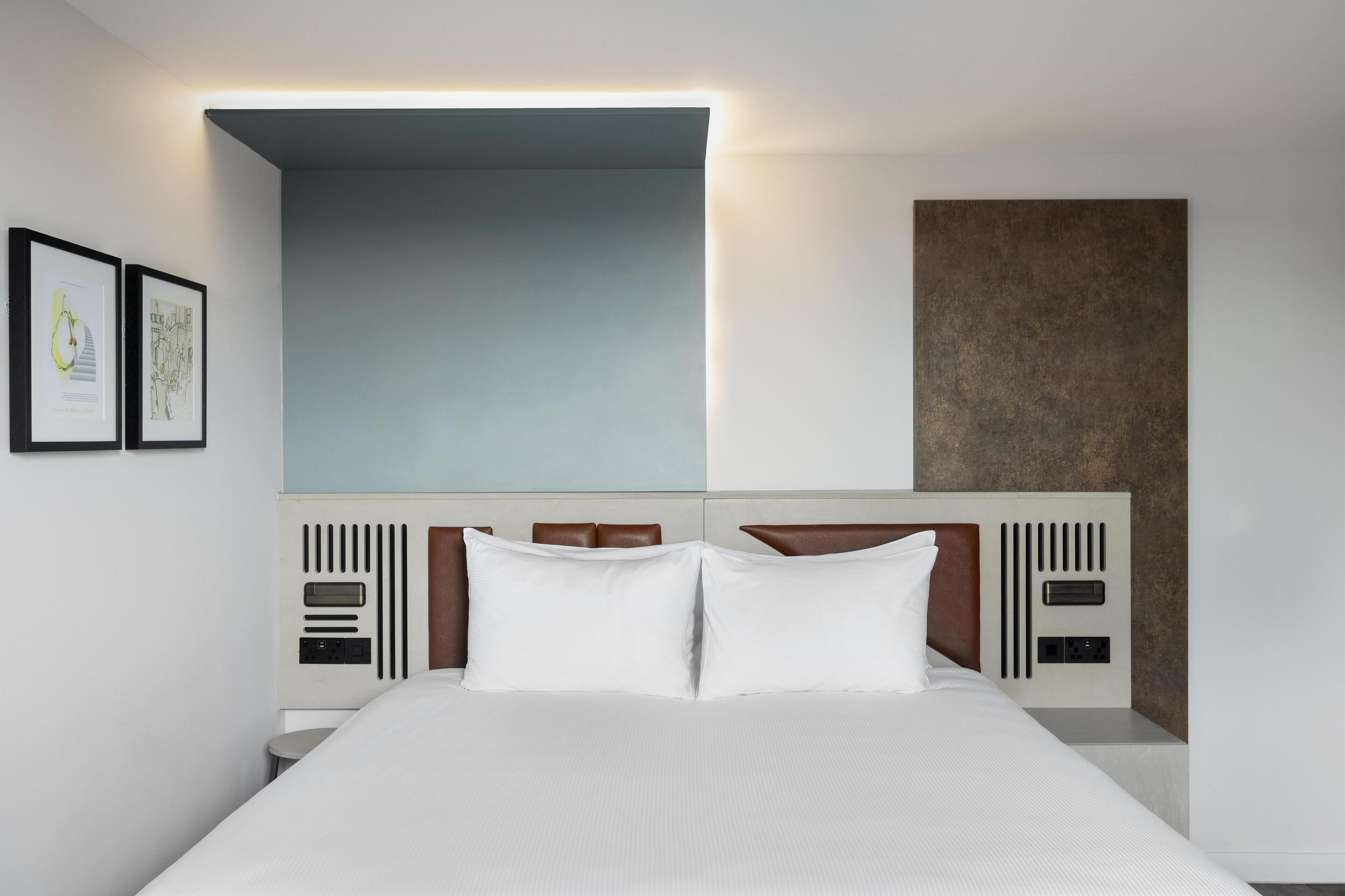 A modern hotel room with a large bed, a headboard with built-in controls, and framed artwork on the wall.