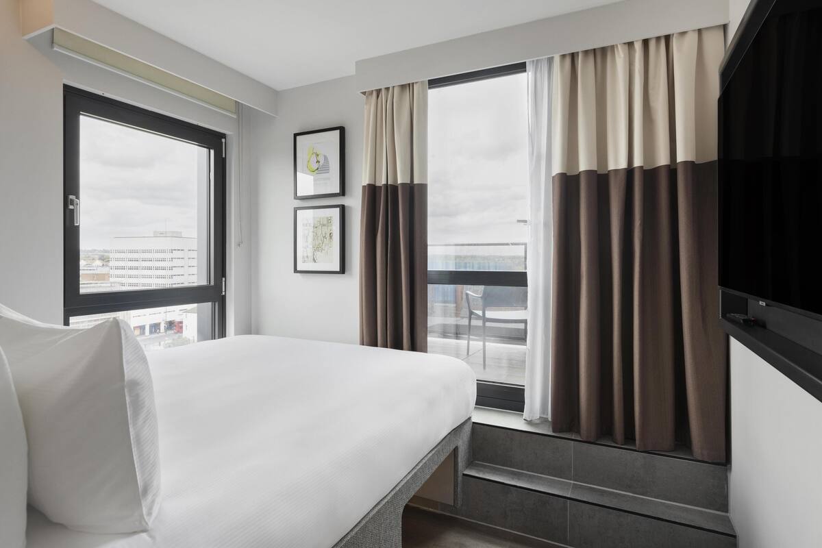 A hotel room with a bed, a window with curtains, a flat-screen TV, and a framed picture on the wall.