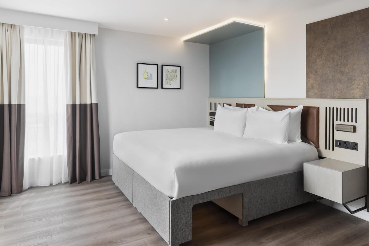 A modern hotel room with a large bed, bedside tables, and a nightstand. There are two framed pictures on the wall, a window with curtains, and a wall-mounted mirror.