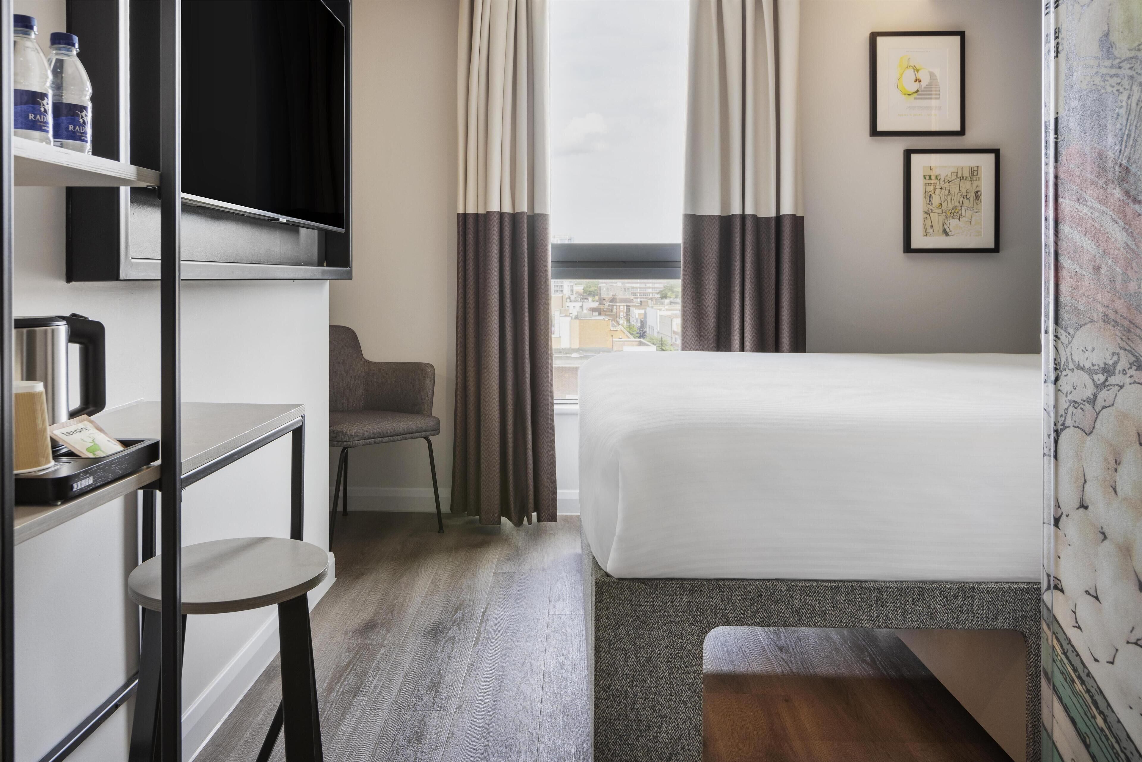 A hotel room with a bed, a desk with a chair, a TV, and a window with curtains.