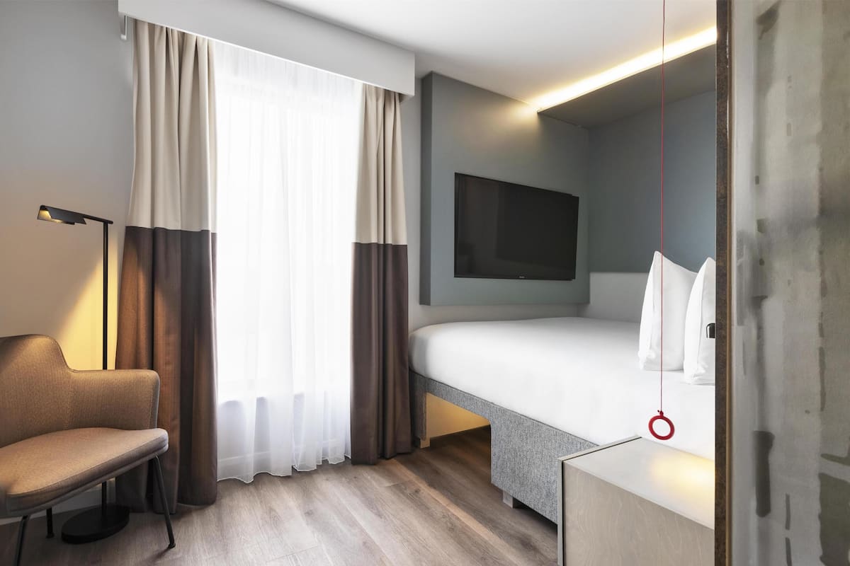 A modern hotel room with a bed, a chair, a TV mounted on the wall, and a window with curtains.