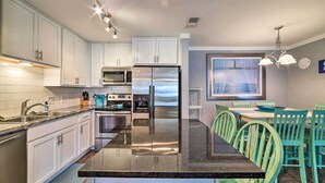 A modern kitchen with white cabinets, stainless steel appliances, a granite countertop, and teal chairs at a dining table. - Pool w/ Spa! Beachfront Condo in Port Aransas (Port Aransas)
